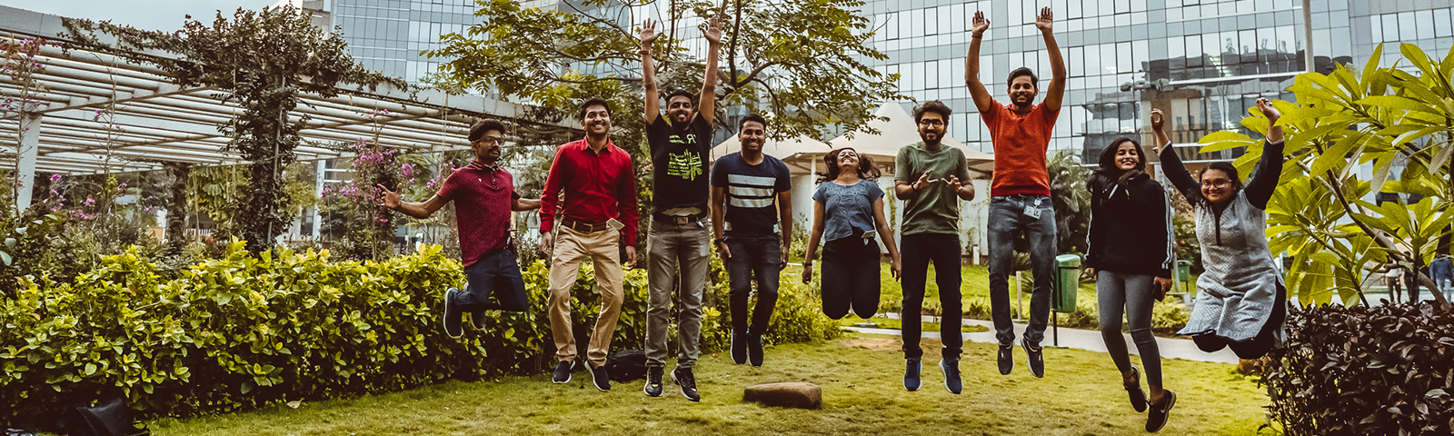 Nine people jumping mid-air outside with a building in the background.