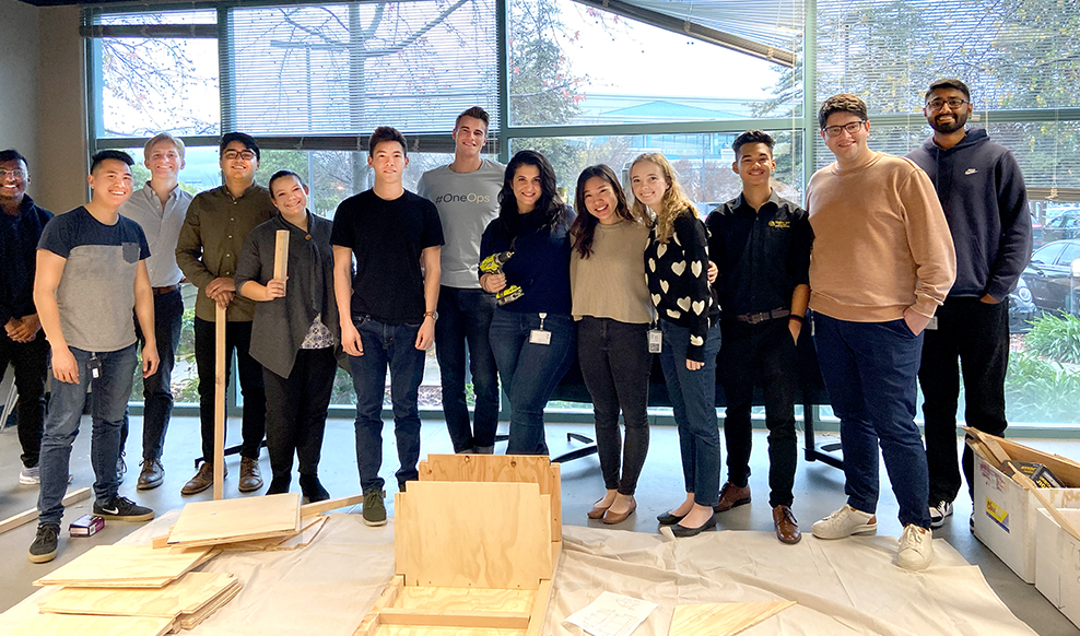 Thirteen employees smiling for a group photo standing in front of wood they are building for volunteer activity