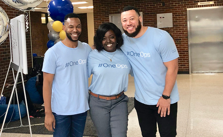 Three people stand together smiling and wearing shirts that read, “#OneOps.”