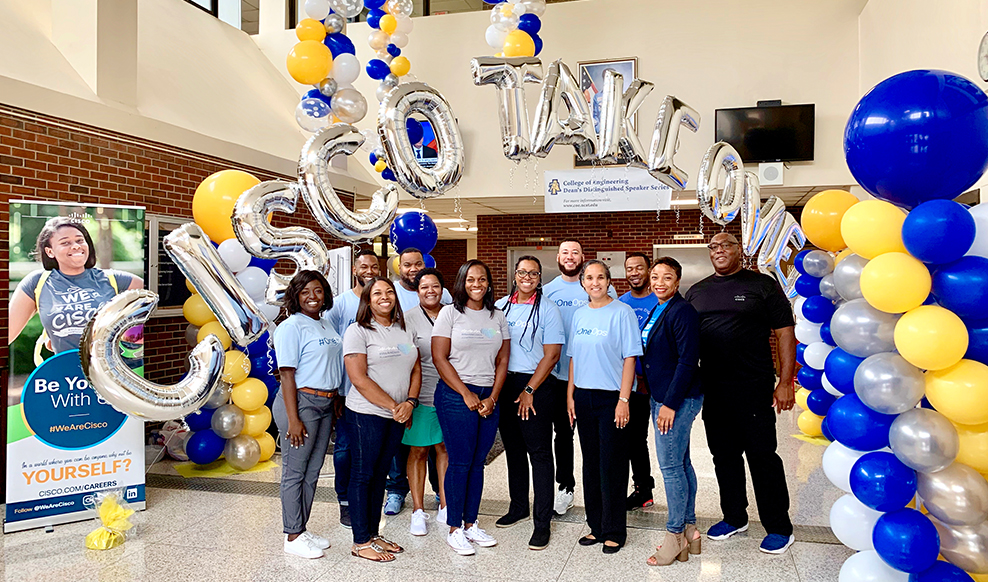 Group of twelve people stand together under balloons that spell out, 'Cisco Takeover.'