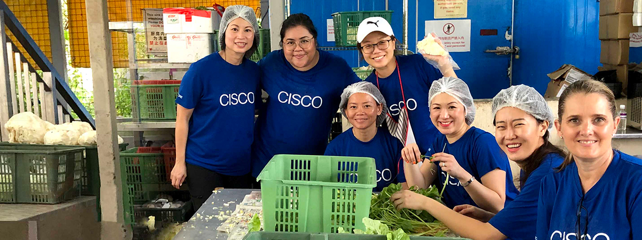 Seven women wearing the same blue Cisco shirt volunteering at a food bank while sorting lettuce