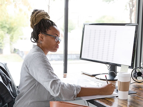 IT professional sitting at a desk with a dashboard on screen and writing notes in a binder.